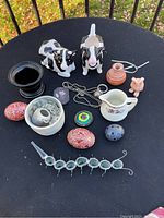 Top-down view of all items arranged on a black table including the cow figurines, decorative eggs, small pitcher, clay pot, rose figurine, black cup, necklace, and candle holder.
