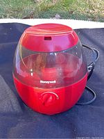 Front angle view of the red Honeywell humidifier sitting outdoors on fabric with power cord visible and water tank clear area.