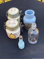 The set of two ceramic preserve jars decorated with pear illustrations, one light blue pottery jar with floral design, one clear glass perfume bottle with gold detailing, and a small blue salt shaker shown on a black surface.