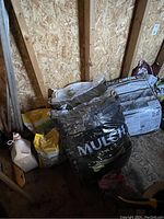 Photo showing several bags of garden supplies including black mulch and soil bags inside a wooden shed.