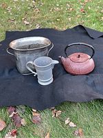 Three metal kitchen items on a black cloth outside on grass: an iron teapot with a black handle, a pewter pitcher, and a wine bucket with handles.