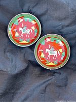 Two round ceramic plates with bright red backgrounds featuring a horseman and colorful figures, set on a dark fabric surface with pewter encasing and plate hangers visible.