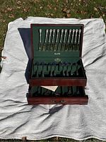Open wood chest displaying multiple Reed & Barton stainless steel knives, forks, and spoons neatly arranged in felt slots inside the chest.
