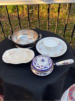 Top view of assorted serving ware on black cloth: silver-tone bowl, oval floral dish, two round floral plates, and decorative saucepan with lid