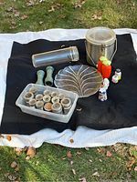 Photo showing entire lot including spice jars, leaf-shaped plate, salt and pepper grinders, glass and metal food storage containers on black cloth outside.