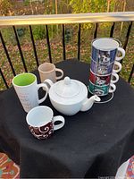 White ceramic teapot and assorted ceramic mugs including stacked animal design mugs with metal holder on black cloth surface.