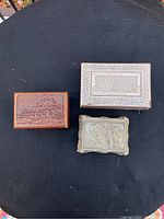 Three closed trinket boxes: a carved wood box, an inlaid wood box, and an ornate metal box on black surface.