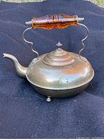 Side angle showing the curved spout, rounded brass body, domed lid, and amber lucite handle attached with metal supports.