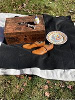 Photo showing wooden box, wooden shoe forms, Quimper ceramic plate, ceramic jar, and ceramic spoon rest arranged outdoors on grass.