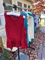 Red sleeveless peplum top followed by various colored and patterned blouses hanging on a rack outdoors.