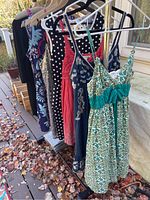 Photo showing six dresses hanging on a rack outdoors, featuring patterns including green-white pattern with teal bow, coral, polka dot black, navy blue with floral, pink floral.