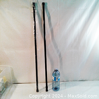 Two long black metal curtain rods standing vertically next to a water bottle for size reference on a white fabric surface