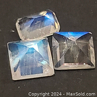 Three faceted genuine natural moonstone gemstones shown resting on a dark surface, displaying their blue-white shimmer and transparency.