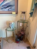 Two gold round side tables with glass tops, large blown glass vase on taller table, ceramic vase on shorter table, and two small decorative vases on floor.