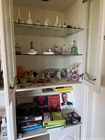 Wide shot of cabinet with upper sections containing porcelain and glass bells and various collectible figurines, lower section showing books and small items.