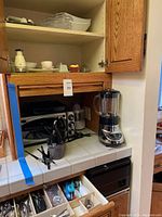 Photo showing a silver Cuisinart toaster oven, a black blender, and a container holding various silverware pieces on tiled kitchen counter and partially opened drawer with utensils.