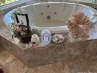Wide view showing all decor items arranged on a corner tub ledge including large coral, basket with flowers, oil burner, photo frame, seashells and shell dish.