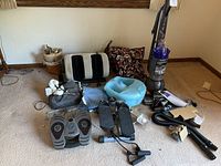 Wide view of the lot showing all items including vacuum, massagers, foot spa, stepper, pillows, and vacuum attachments on carpeted floor.