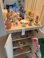 Wide view of countertop and open cabinet filled with various decorative glassware and ceramic items including vases, figurines, small clock, and candle holders.