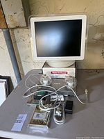 Photo of the vintage CRT iMac monitor on top of a Maxtor OneTouch II box with Canon device, dual glass lamps, power strip, and cables laying on table.