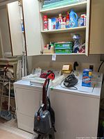 Photo showing white washer and dryer with multiple cleaning products on shelves above, and a Hoover vacuum cleaner in front.