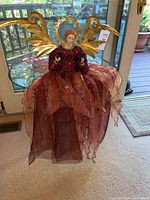 Angel figurine with flowing burgundy and pink fabric dress and gold wings placed on carpet in front of door.