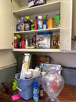 White shelves holding various cleaning products such as sprays, liquids, and wipes. Blue bucket and clear glass vase on work surface.