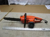Side view of the orange Remington Mity Mite electric chainsaw showing the full 12-inch cutting bar and power cord, placed next to measuring tape for size reference.