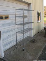 Five shelf steel wire rolling rack standing outside in front of a closed garage door. The wire shelves are evenly spaced and mounted on tubular posts. Four swivel caster wheels attached for mobility.