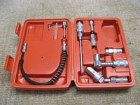Photo of the 9 piece greasing accessory kit in its open orange plastic carrying case showing eight metal accessories: straight and swivel grease couplers and a flexible hose with spring guard.