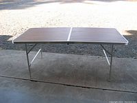 Clear image of the aluminum folding table fully set up outdoors, showing the full surface with brown wood-grain pattern and aluminum edging