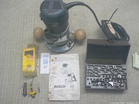 Bosch router with wooden handles beside router bits in plastic case and instruction manual on carpeted floor
