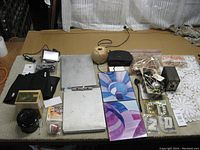 Wide view of entire lot showing camera, sewing tools, clothespins, string, picture frame, document holders, knee braces, clock, crochet piece, and candle snuffer arranged on a table.