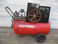 Side view of Craftsman portable air compressor showing red tank, black motor housing, wheels, pressure gauges, and handle.