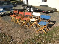 Five folding directors' chairs lined up outdoors on gravel and grass. Wooden frames with canvas seats and backs in red, off-white, and blue. Visible wear and soiling on canvas. Two chairs show 'Telescope' branding on canvas.