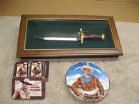 A display showing the John Wayne commemorative Bowie cutter knife in a wooden and glass case, two decks of John Wayne playing cards in a metal tin, and the Franklin Mint collector plate with John Wayne's image.