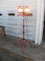 Full view of the large red tripod mounted dual head halogen work light illuminated outdoors near garage door.