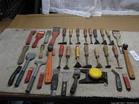 Photo showing approximately 35 paint scrapers of various shapes, sizes, and handle materials laid out on fabric in used condition