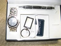Photo of the watch, pen, keychain, and branded card inside white molded presentation insert in black box with loose lid.