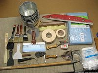 Photo showing all the metal paint buckets stacked with various paint brushes, masking tapes, corner edgers, spackle tools, trowels, sanding pole, and a blue disposable drop cloth folded.