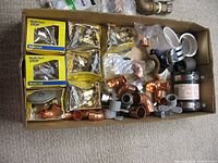 Box containing BrassCraft Multi-Turn Stop valves in original packaging, various loose copper fittings, PVC pipe fittings, and white flange covers.
