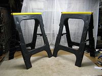 Pair of black plastic folding sawhorses with yellow top edges shown side by side, standing open on carpeted floor against a fabric backdrop.
