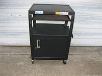 Front view of a black metal rolling media cart showing three levels: top flat surface, sliding middle tray, and enclosed lower cabinet with a latch door. Four caster wheels visible.