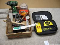 Photo showing an array of tools including a green Hitachi impact wrench, drill bits, wire brush, folding carpenter rulers, combination square, stud finder in case, tubing bender, and other hand tools arranged in a box and on the floor.