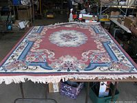 Full view of rectangular area rug spread on table, showing pink background with floral and scroll pattern in blue, white, black and pink hues, with fringe edges.