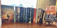 Photo of a row of assorted hardcover and paperback books including titles such as 'Fives and Twenty-Fives' by Michael Pitre and 'To Die Beautiful' by Buzzy Jackson.