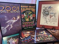 Five framed Mardi Gras Galveston posters displayed on a wooden surface with easels, representing event years 2000, 2001, 2002, 2003, and 2005.