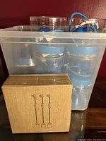 Multiple blue 1-light pendant hanging lights inside a clear plastic bin alongside a cardboard box showing packaging details.