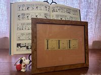 Photo showing a framed Peanuts comic strip with Snoopy and Woodstock visible, wood-tone frame, and a Peanuts Treasury book open in the background.