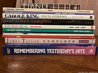Stack of 10 music books featuring piano, vocal, and guitar material. Titles include Debussy, Carole King, Paul Simon, Christmas songs, Beatles, James Taylor, and Remembering Yesterday's Hits.